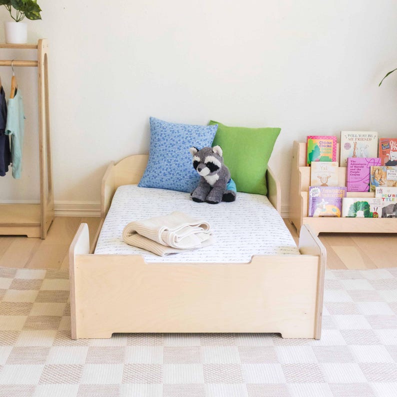 May include: A light wood toddler bed with a mattress, pillows, and a blanket. A stuffed raccoon sits on the bed. A wooden bookshelf with children's books is to the right. A wooden clothing rack is to the left.