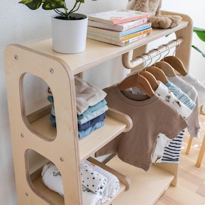K&ouml;nnte beinhalten: Ein Holz-Kleiderst&auml;nder mit Regalen zur Aufbewahrung. Der Kleiderst&auml;nder hat eine nat&uuml;rliche Holzoptik und verf&uuml;gt &uuml;ber eine Kleiderstange mit Holzb&uuml;geln. Die Regale sind mit gefalteten Kleidungsst&uuml;cken gef&uuml;llt, darunter ein Stapel blauer und gr&uuml;ner Hemden und ein Stapel wei&szlig;er und schwarzer gemusterter Babydecken.
