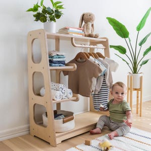 K&ouml;nnte beinhalten: Ein Kinderkleiderschrank aus Holz mit Regalen, einer Kleiderstange und einem Korb. Ein Kind sitzt vor dem Schrank und tr&auml;gt ein gr&uuml;nes Shirt und eine graue Hose. Der Schrank ist aus hellem Holz und hat eine nat&uuml;rliche Oberfl&auml;che.