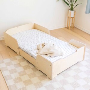 May include: A light-coloured wooden toddler bed with a patterned sheet. A beige blanket and soft toy rest on the bed. The bed is placed on a checkered rug. The bed's dimensions are approximately 140 cm in length.