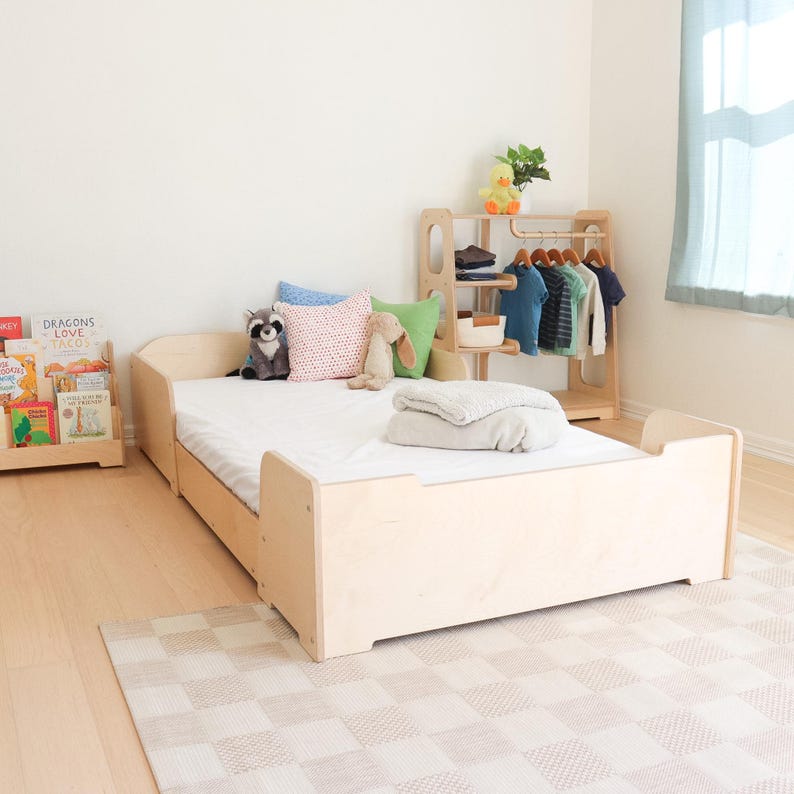 May include: A light-coloured wooden toddler bed with a white mattress, pillows, and a blanket. A small wooden clothing rack with colourful shirts and a bookshelf with children's books are also in the room. A stuffed raccoon and bunny are on the bed.