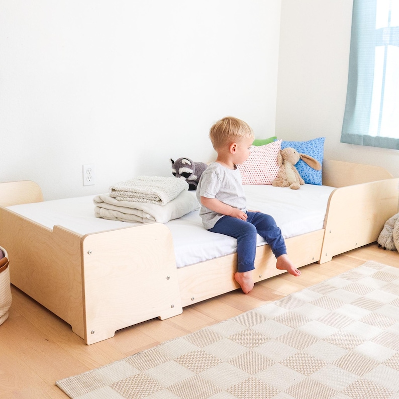 May include: A light-coloured wooden toddler bed with a white mattress. A young child sits on the bed. Pillows and a folded blanket are on the bed. A stuffed raccoon and a stuffed bunny are also on the bed.