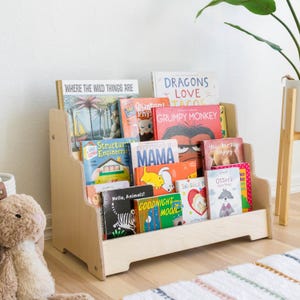 May include: A wooden bookshelf with three shelves, filled with colorful children's books. The books have titles like "Where the Wild Things Are", "Dragons Love Tacos", "Grumpy Monkey", and "Everything is Mama".