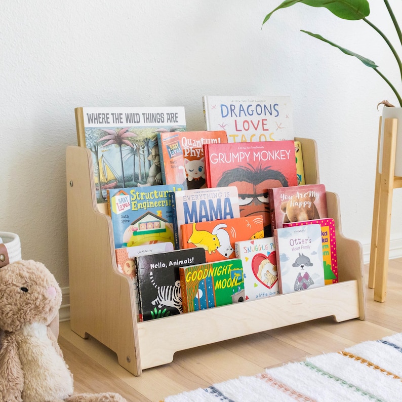 May include: A wooden bookshelf with three shelves, filled with colorful children's books. The books have titles like "Where the Wild Things Are", "Dragons Love Tacos", "Grumpy Monkey", and "Everything is Mama".