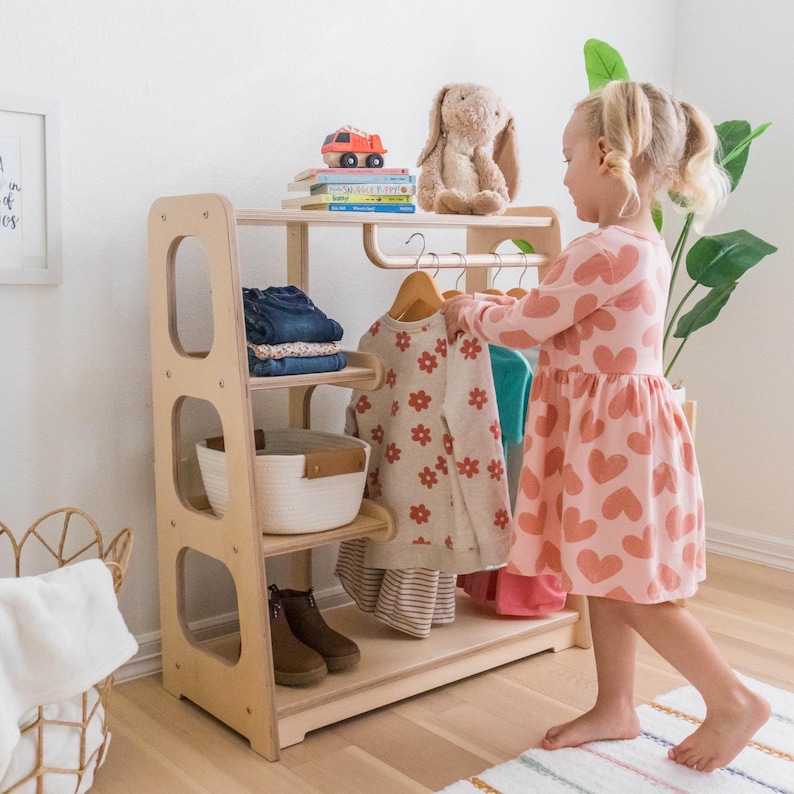 Young Girl hanging her clothes on a Montessori wardrobe built at the height for young kids. The wardrobe is made from natural wood.