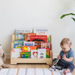 May include: A wooden bookshelf with a variety of children's books, including "Where the Wild Things Are", "Dragons Love Tacos", and "Grumpy Monkey". A young child is sitting on the floor in front of the bookshelf, reading a book.