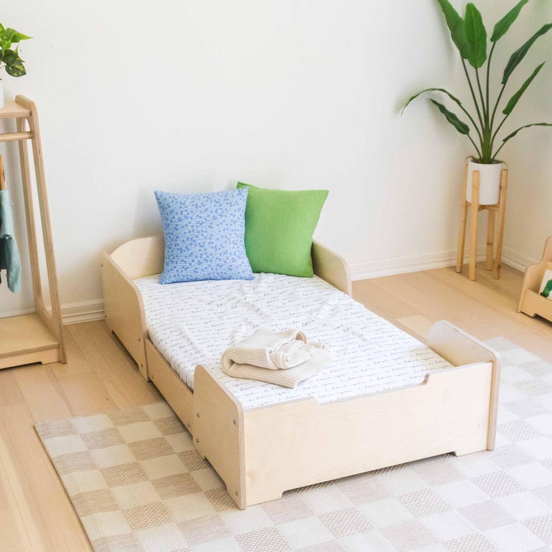 May include: A light wood toddler bed with a mattress, two pillows, and a folded blanket. The bed has a low profile with rounded edges and a simple design. A blue floral pillow and a green pillow are on the bed.