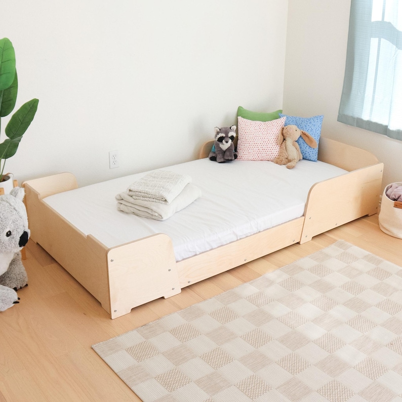 May include: A light-coloured wooden toddler bed with a white mattress, designed with rounded edges for safety. The bed is accessorised with pillows, a stuffed raccoon, a stuffed bunny, and a folded blanket. A neutral rug is on the floor.