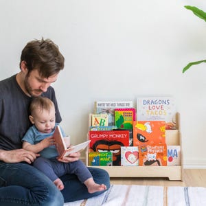 May include: A wooden bookshelf with multiple shelves holds a variety of colorful children's books. The books include titles like "Where the Wild Things Are", "Chicka Chicka Boom Boom", "Dragons Love Tacos", "Grumpy Monkey", and "Goodnight Moon". A father and son are sitting on the floor in front of the bookshelf, reading a book together.