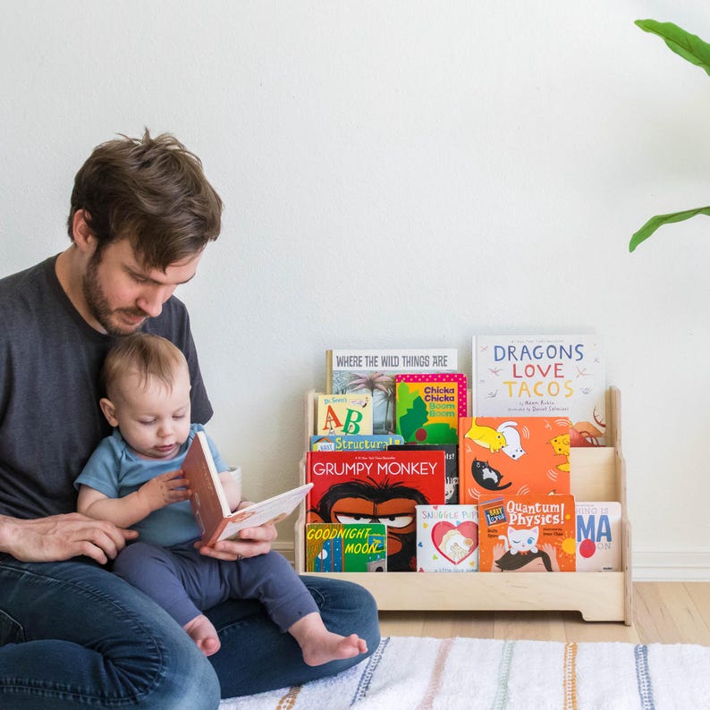 May include: A wooden bookshelf with multiple shelves holds a variety of colorful children's books. The books include titles like "Where the Wild Things Are", "Chicka Chicka Boom Boom", "Dragons Love Tacos", "Grumpy Monkey", and "Goodnight Moon". A father and son are sitting on the floor in front of the bookshelf, reading a book together.
