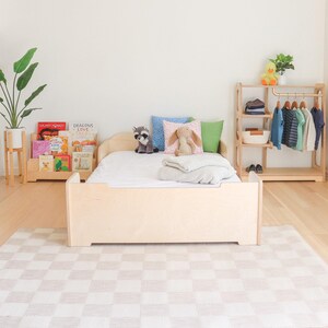 May include: A light-coloured wooden toddler bed with a low profile, featuring a white mattress and pillows. A wooden bookshelf with children's books and a wooden clothing rack with hanging clothes are also visible. A checkered rug covers the floor.