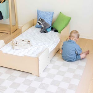 May include: A light wood toddler bed with a patterned mattress, a blue cushion, and a green cushion. A stuffed raccoon sits on the bed. A folded blanket rests on the mattress. A child sits on the floor near the bed.