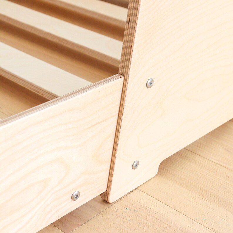 May include: Close-up of a wooden bed frame with visible wood grain. The frame is constructed with light-coloured wood and features a slatted base. Metal screws secure the frame's panels. The bed frame is on a light-coloured wooden floor.