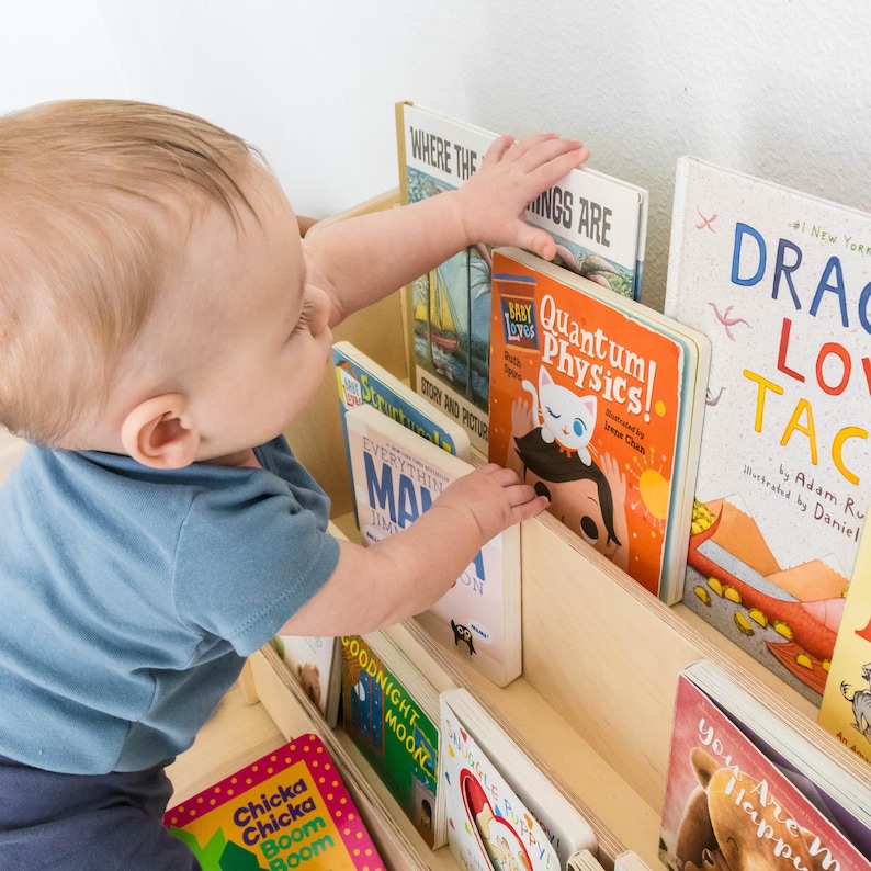 May include: A child reaches for a book titled "Quantum Physics!" on a wooden bookshelf filled with colorful children's books. The bookshelf has two shelves and is filled with a variety of books.