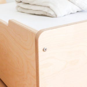 May include: Close-up of a light-coloured wooden bed frame with a white top surface. The frame has rounded corners and is assembled with a silver bolt. A folded white blanket rests on the bed.