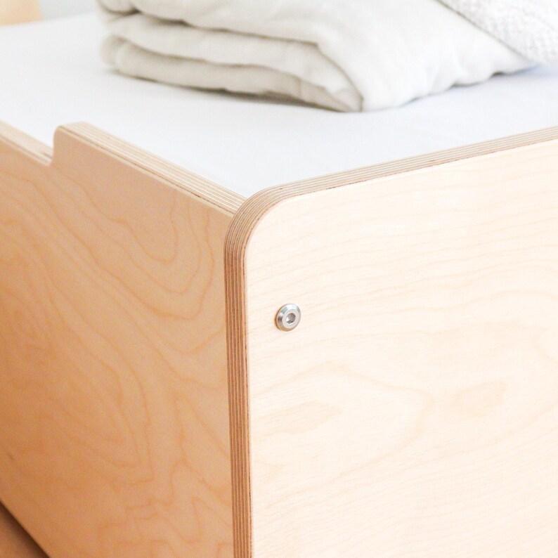 May include: Close-up of a light-coloured wooden bed frame with a white top surface. The frame has rounded corners and is assembled with a silver bolt. A folded white blanket rests on the bed.