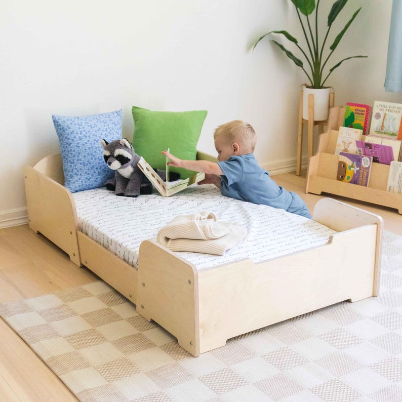 May include: A light wood toddler bed with a mattress, pillows, and a blanket. A child is reaching for a book on the bed. A stuffed raccoon and a green pillow are on the bed. A bookshelf with books is in the background.