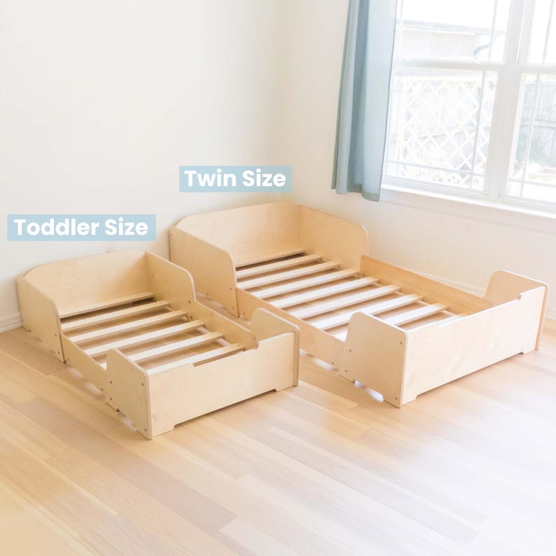 May include: Two wooden toddler beds, one labelled "Toddler Size" and the other "Twin Size." Both beds have a natural wood finish, slatted bed frames, and low side rails for safety. The beds are designed for children.