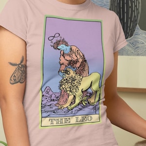 May include: A pink t-shirt with a graphic of a tarot card depicting The Leo. The card features a woman with flowing hair and a lion. The text "THE LEO" is printed at the bottom of the card.