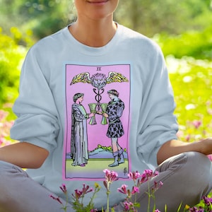 May include: A person wearing a light blue sweatshirt with a pink and blue tarot card design featuring the Two of Cups. The card depicts two figures holding cups, with a winged serpent above them. The person is sitting in a field of pink flowers with their eyes closed.