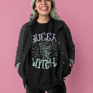 May include: A black t-shirt with a graphic print of a snake and the text "Queer Witch" in a pastel purple and blue color scheme. The person wearing the shirt is smiling and wearing a gray beanie.