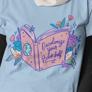 May include: A light blue t-shirt with a graphic of an open book with flowers and the text "Decolonize your Bookshelf".