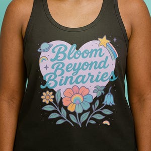 May include: Dark gray tank top with a heart-shaped graphic that reads "Bloom Beyond Binaries." The design includes a planet, star, rainbow, and flowers in pastel colors. The tank top is worn by a person with dreadlocks and glasses.