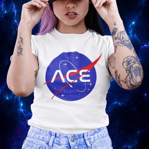 May include: White t-shirt with a blue circle logo featuring the word "ACE" in white, with a red swoosh. The shirt is paired with light blue jeans and a black baseball cap. The background is a dark blue space scene.