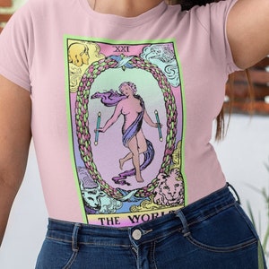 Op de afbeelding: Een roze T-shirt met een kleurrijke illustratie van de tarotkaart Wereld. De kaart toont een vrouw die in een cirkel staat, omringd door een leeuw, een stier, een engel en een adelaar. De tekst "XXI DE WERELD" is onder de kaart gedrukt.