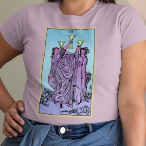 Pode incluir: Uma mulher vestindo uma camiseta lavanda com um design de carta de tarô que apresenta três figuras segurando taças. O cartão é intitulado "III" e pertence ao naipe de Copas. A mulher também está vestindo jeans azul e uma jaqueta de jeans amarrada na cintura.