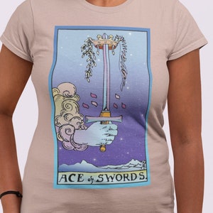 May include: A beige t-shirt with a graphic of the Ace of Swords tarot card. The card features a blue and purple background with a hand holding a sword.