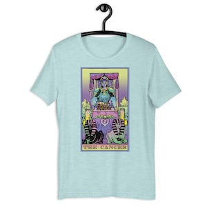 Puede incluir: Una camiseta azul claro con una ilustraci&oacute;n colorida de la carta del tarot El C&aacute;ncer. La carta presenta una figura con una corona y armadura sentada en un trono, con dos figuras parecidas a esfinges en la parte inferior. El texto "THE CANCER" est&aacute; impreso debajo de la imagen.