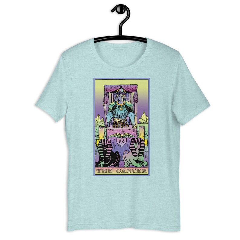 Puede incluir: Una camiseta azul claro con una ilustraci&oacute;n colorida de la carta del tarot El C&aacute;ncer. La carta presenta una figura con una corona y armadura sentada en un trono, con dos figuras parecidas a esfinges en la parte inferior. El texto "THE CANCER" est&aacute; impreso debajo de la imagen.