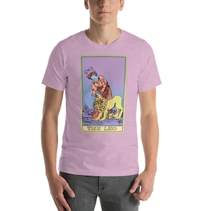 May include: A pink t-shirt with a graphic of a tarot card, The Leo, featuring a person with a halo and a lion. The card has a purple, blue, and yellow gradient background.