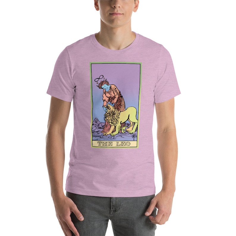 May include: A pink t-shirt with a graphic of a tarot card, The Leo, featuring a person with a halo and a lion. The card has a purple, blue, and yellow gradient background.