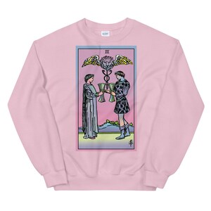 May include: Pink sweatshirt with a colorful illustration of the Two of Cups tarot card. The card features two figures holding cups, a winged serpent, and the Roman numeral II.