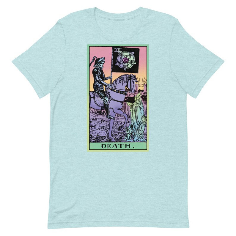 Puede incluir: Camiseta azul claro con un dise&ntilde;o de carta del tarot. El gr&aacute;fico representa una figura esquel&eacute;tica a caballo, sosteniendo una bandera con una rosa. La palabra "DEATH" est&aacute; impresa debajo. El dise&ntilde;o est&aacute; enmarcado en un borde rosa y verde.