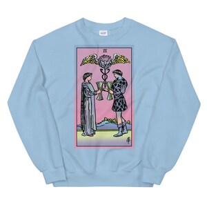May include: Light blue sweatshirt with a graphic of the Two of Cups tarot card. The card features two figures in robes holding cups, with a winged serpent and a lion's head above them. The card is surrounded by a pink and green border.