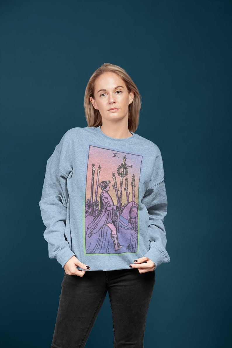 May include: A light blue sweatshirt with a colorful tarot card design featuring the Six of Wands. The card shows a rider on horseback with a wreath of flowers on a staff.