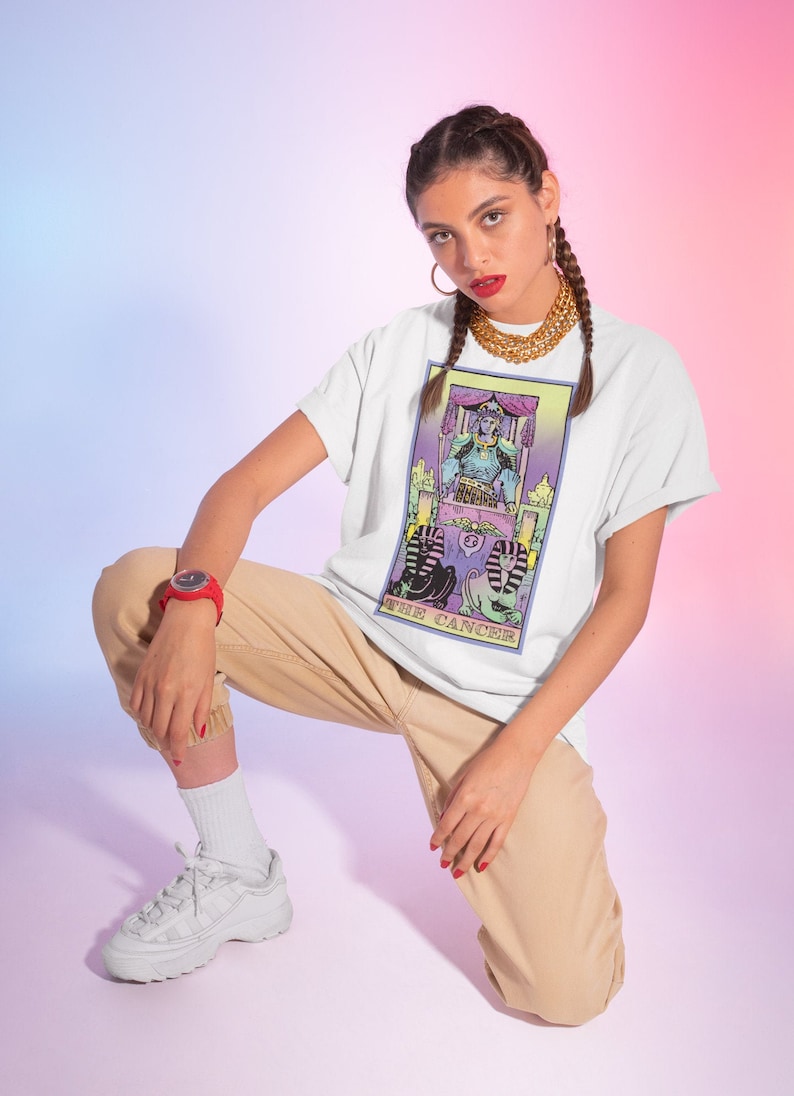 Puede incluir: Camiseta blanca con un dise&ntilde;o de carta de tarot colorido que presenta el texto "The Cancer". La carta representa una figura sentada en un trono con dos figuras de esfinge en la parte inferior.