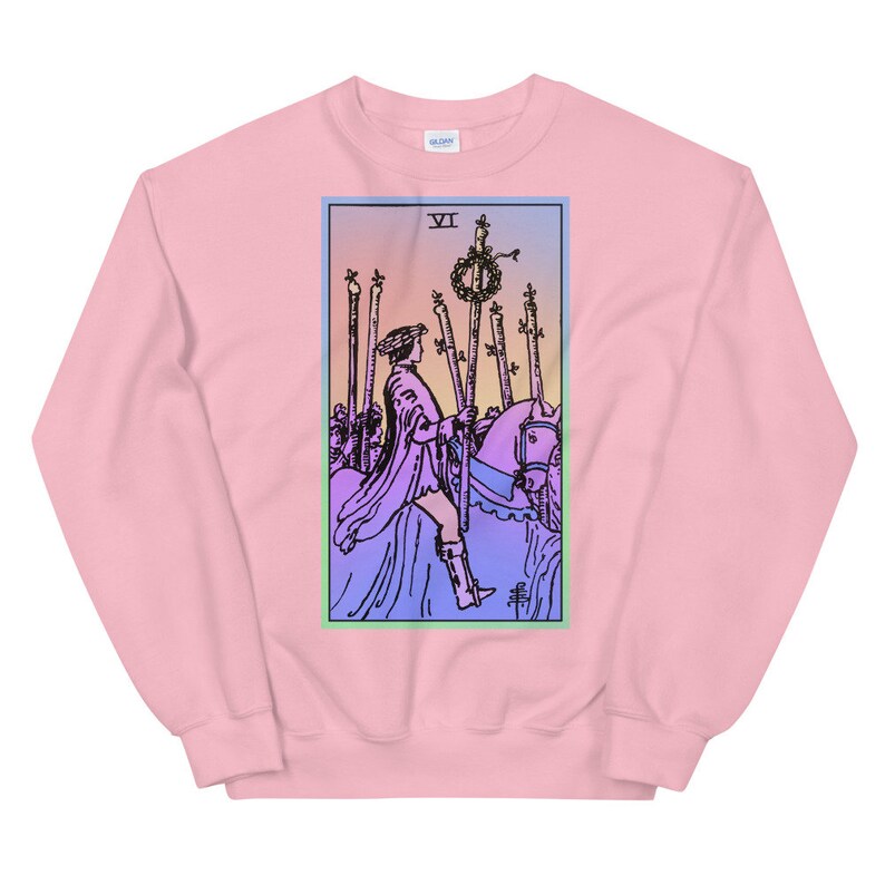 May include: Pink sweatshirt with a colorful illustration of the Six of Wands tarot card. The card features a rider on horseback, holding a staff with a wreath on top. The card is surrounded by a light blue, green, and pink border.