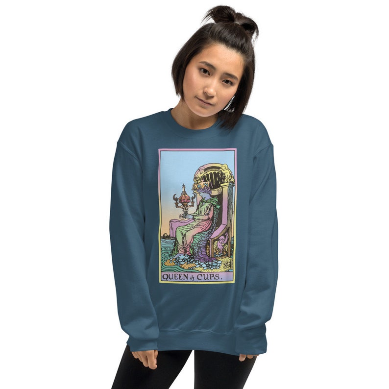 Puede incluir: Una sudadera azul turquesa con una ilustraci&oacute;n colorida de la carta del tarot de la Reina de Copas. La carta muestra a una mujer sentada en un trono con una copa en la mano. El texto "QUEEN of CUPS." est&aacute; impreso debajo de la imagen.