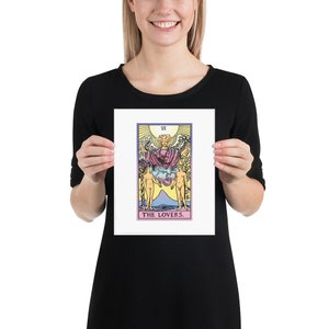 May include: A tarot card print featuring the Lovers card from the Rider-Waite deck. The card depicts a winged angel with a crown above a couple standing on either side of a mountain. The card is framed in a white border with the Roman numeral VI and the words "THE LOVERS."