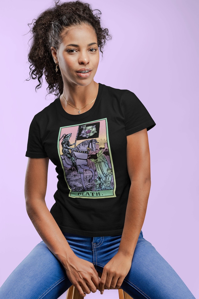 Puede incluir: Camiseta negra con un dise&ntilde;o de carta del tarot que representa la Muerte. El dise&ntilde;o muestra una figura con t&uacute;nica a caballo, sosteniendo una bandera, con la palabra "DEATH" debajo. El gr&aacute;fico tiene colores pastel y est&aacute; enmarcado.