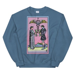 May include: A blue sweatshirt with a graphic print of the Two of Cups tarot card. The card features two figures holding cups, a winged serpent, and a lion's head.