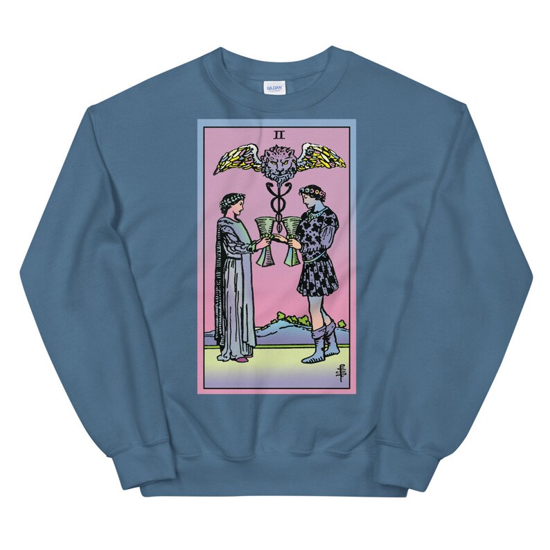 May include: A blue sweatshirt with a graphic print of the Two of Cups tarot card. The card features two figures holding cups, a winged serpent, and a lion's head.