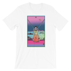 Op de afbeelding: Wit T-shirt met een kleurrijke illustratie van de tarotkaart Twee van Zwaarden. De kaart toont een persoon die op een troon zit, met een blinddoek om en twee gekruiste zwaarden vasthoudt. De achtergrond is een zeelandschap met een halve maan aan de hemel.