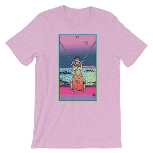 Op de afbeelding: Een roze T-shirt met een afbeelding van de tarotkaart Twee van Zwaarden. De kaart toont een persoon die op een troon zit met hun ogen bedekt door twee zwaarden. De achtergrond is een verloop van roze, blauw en groen.