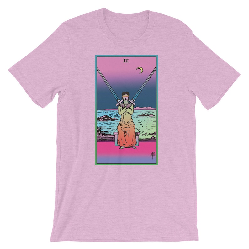 Op de afbeelding: Een roze T-shirt met een afbeelding van de tarotkaart Twee van Zwaarden. De kaart toont een persoon die op een troon zit met hun ogen bedekt door twee zwaarden. De achtergrond is een verloop van roze, blauw en groen.