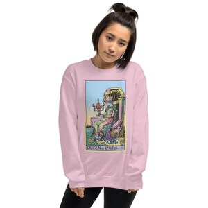 Puede incluir: Sudadera rosa con un gr&aacute;fico de la carta del tarot de la Reina de Copas. La carta presenta a una mujer sentada en un trono con una copa en la mano. El texto "QUEEN of CUPS." est&aacute; impreso debajo de la carta.
